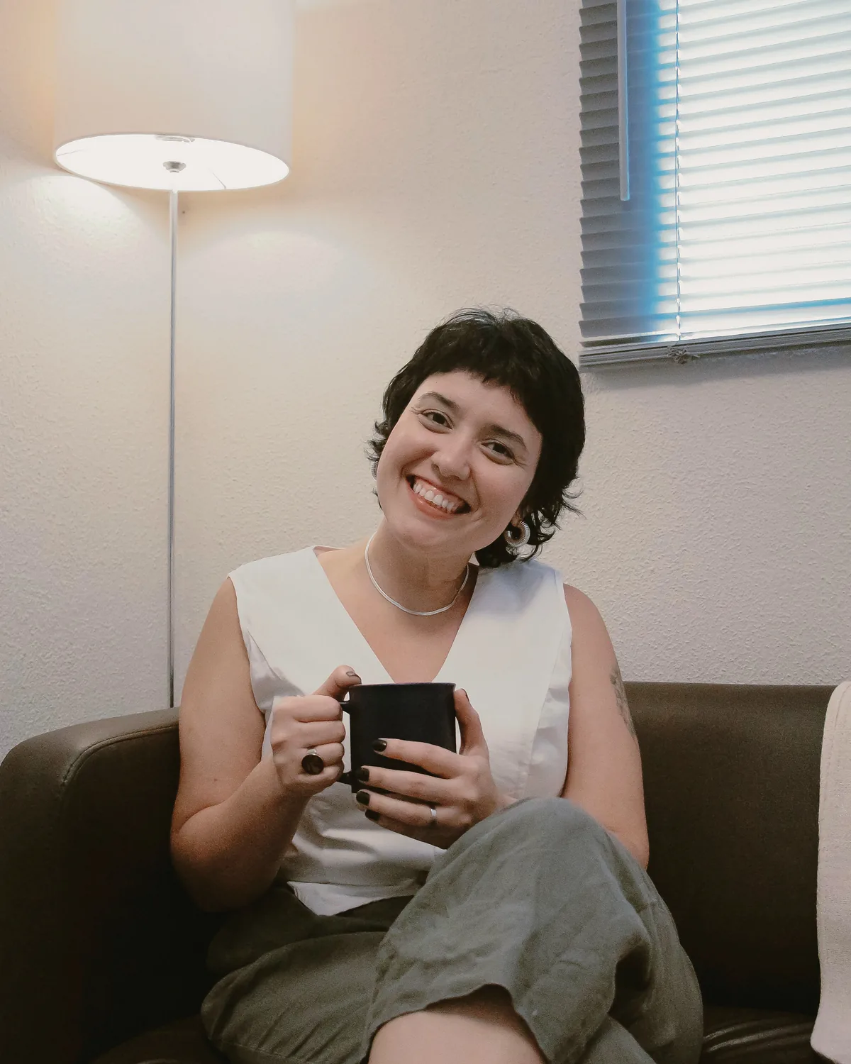 Veri sorrindo com uma caneca, sentada num sofá ao lado de um abajur