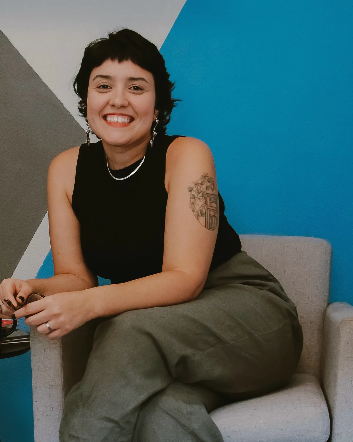 Veri sorrindo no consultório, sentada numa poltrona clara diante de uma parede pintada em cinza e azul